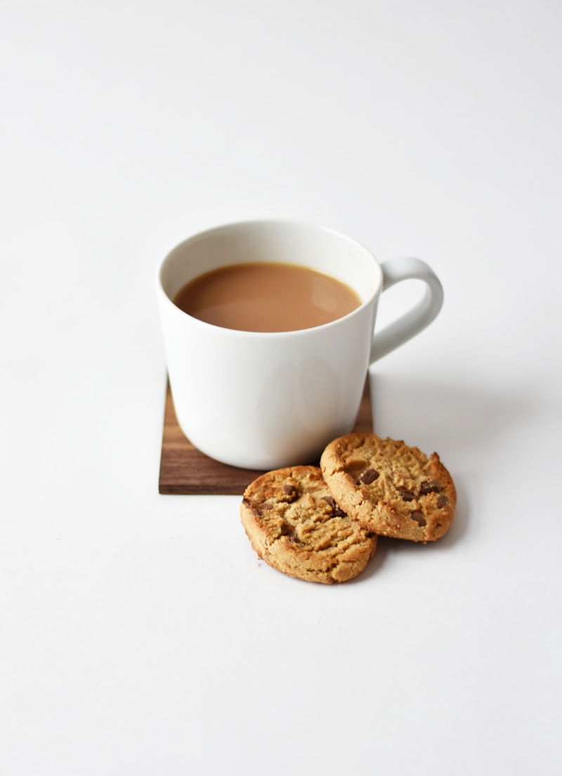 White mug of coffee with milk beside two cookies on a wooden coaster